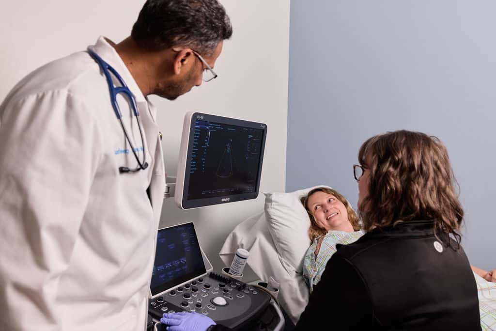 Doctor and ultrasound sonographer reviewing imaging scan results with a patient lying on an exam bed during a clinical consultation for Carotid Artery Disease Treatment