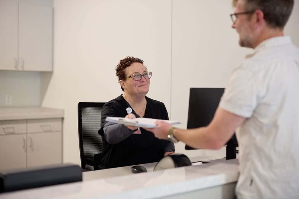 Clinic receptionist handing intake paperwork on a clipboard to a patient at the front check-in desk for Conditions We Treat