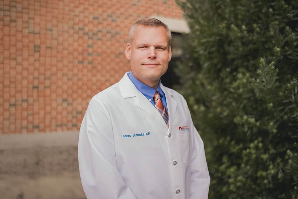 Nurse practitioner smiling outdoors in a white lab coat for a professional provider portrait of Marc Arnold