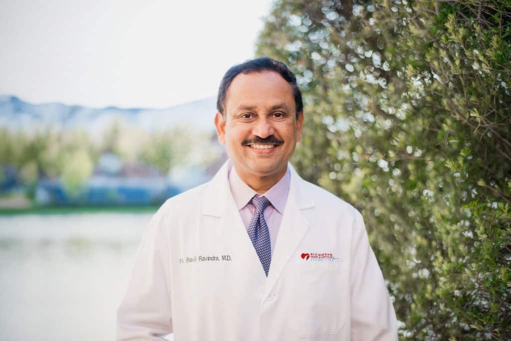Board-certified cardiologist smiling in a white lab coat outdoors for a professional provider portrait of P. V. Ravindra, MD, FACC